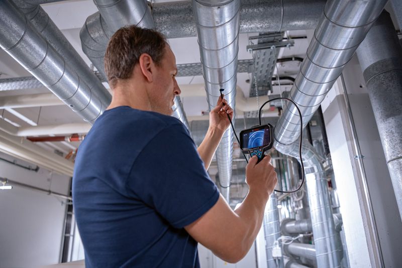 A person inspects overhead pipes using an inspection camera in an industrial setting.