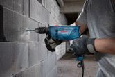 A person wearing safety equipment drills into a concrete block wall with an impact drill.