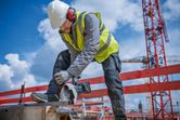 A person wearing safety equipment uses a cordless angle grinder to cut metal at a construction site.