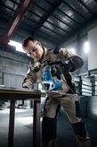 A person wearing safety equipment uses a jigsaw to cut metal in a workshop.