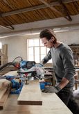 A person wearing safety equipment cuts wood with a mitre saw in a workshop.