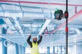A person wearing safety equipment aligns a ceiling panel using a laser leveling tool on a telescopic pole.