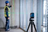 Person wearing safety equipment aligns framing studs using a laser leveling tool on a tripod.