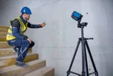 A person wearing safety equipment marks a wall using a laser leveling tool on a tripod.