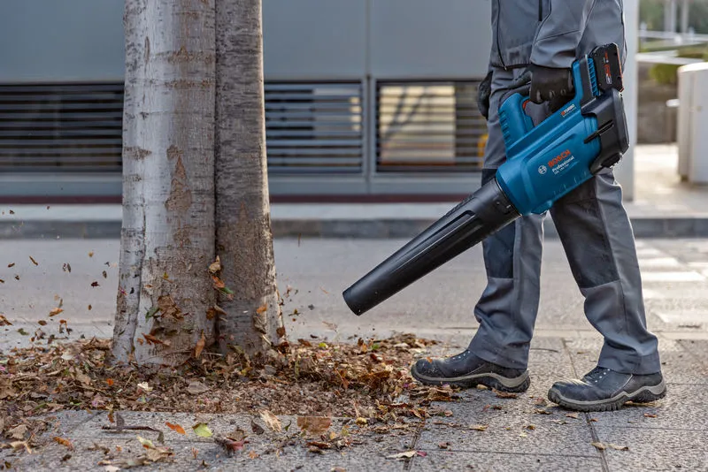GBL 18V-750 A person wearing safety equipment uses a cordless leaf blower to clear leaves near a tree.