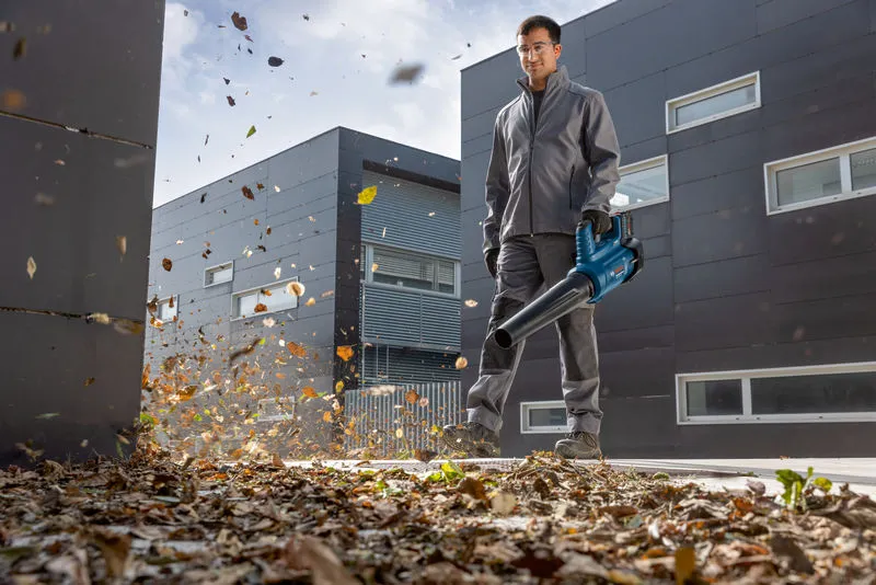 GBL 18V-750 A person wearing safety equipment clears leaves with a cordless leaf blower outside a building.