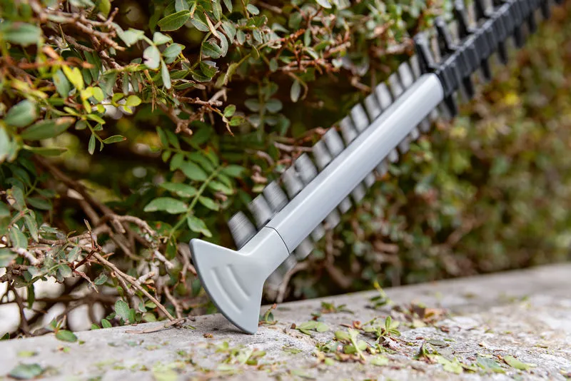 GHE 18V-60 Hedge trimmer blade cutting green shrubbery along a garden edge.