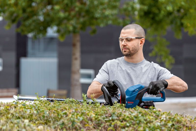 GHE 18V-60 A person wearing safety equipment trims bushes using a cordless hedge trimmer.
