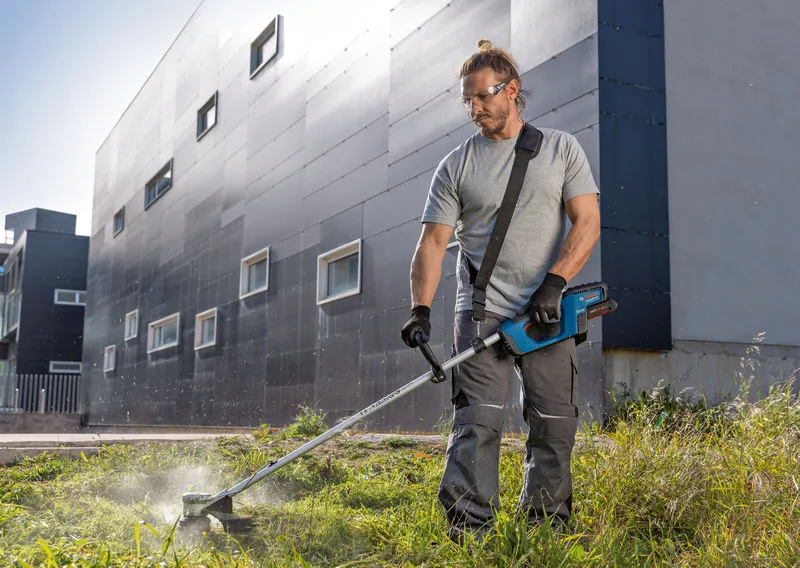 GRT 18V-33 A person wearing safety equipment trims grass with a cordless grass trimmer outside a building.
