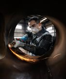 A person wearing safety equipment grinds metal inside a large pipe, creating sparks.