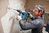 A person wearing safety equipment uses a rotary hammer to remove tiles from a wall.