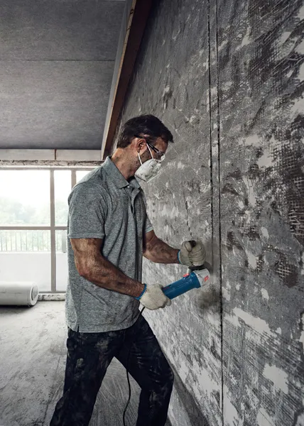 A person wearing safety equipment uses an angle grinder to cut into a concrete wall.