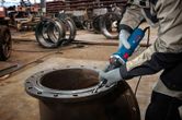 A person wearing safety equipment grinds the edge of a large metal pipe with a straight grinder.