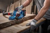 A person wearing safety equipment planes a wooden plank with a cordless tool.