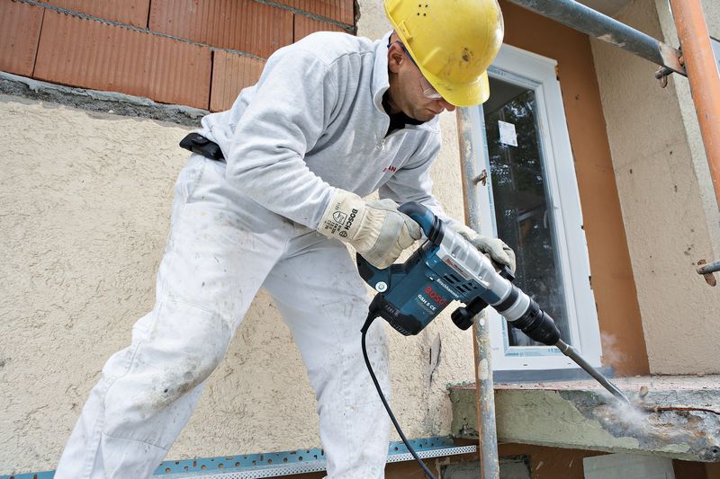 A person wearing safety equipment uses a rotary hammer to break concrete on a building site.