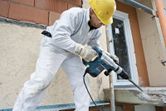 A person wearing safety equipment uses a rotary hammer to break concrete on a building site.