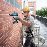 A person wearing safety equipment drills into a brick wall on scaffolding.