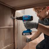 A person wearing safety equipment uses a cordless drill to install a cabinet hinge.