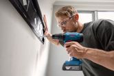 A person wearing safety equipment drills into a wall to install a mounting bracket.