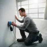 A person wearing safety equipment sands a wall with a power sander.
