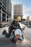 A person wearing safety equipment operates a metal cut-off saw, producing sparks on steel beams.