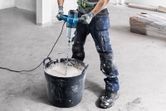 A person wearing safety equipment mixes paint with an electric stirring mechanism.