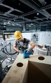 A person wearing safety equipment uses a jigsaw to cut wood at a construction site.