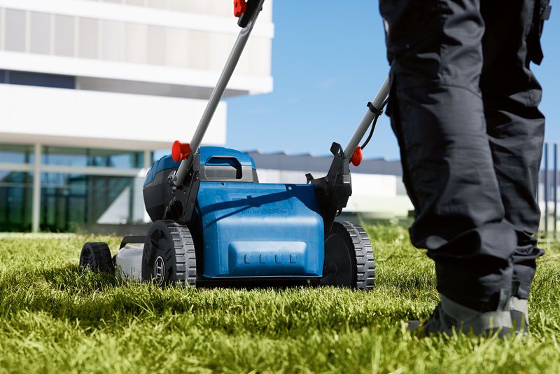 A person wearing safety equipment pushes a cordless lawn mower on green grass.