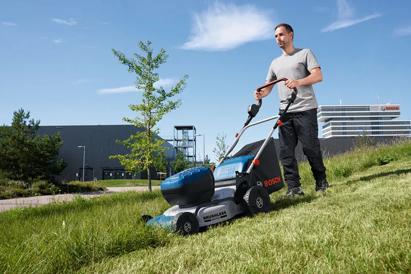 A person pushes a cordless lawnmower across a grassy slope outdoors.