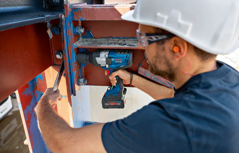 A person wearing safety equipment uses a cordless impact wrench on a steel beam.