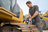 A person wearing safety equipment uses a cordless impact wrench on an excavator track.