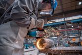 A person wearing safety equipment grinds a metal pipe with an angle grinder, sparks flying.