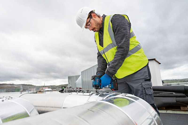 A person wearing safety equipment uses a power drill on a large metal duct outdoors.
