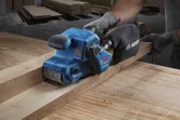 A person wearing safety equipment sands a wooden plank with a blue belt sander.