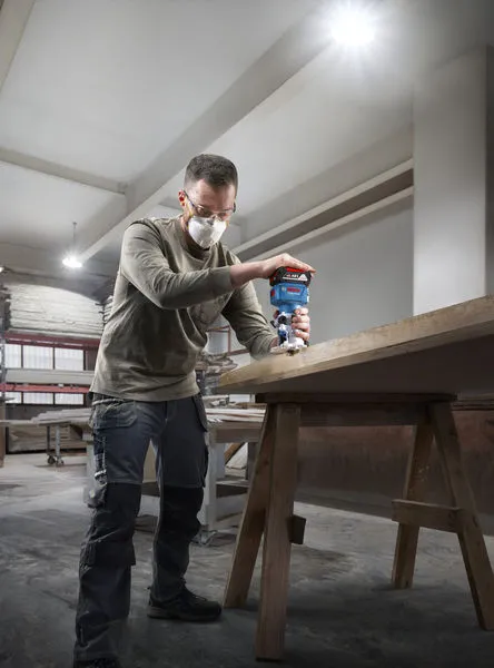 A person wearing safety equipment uses a router on the edge of a wooden table.