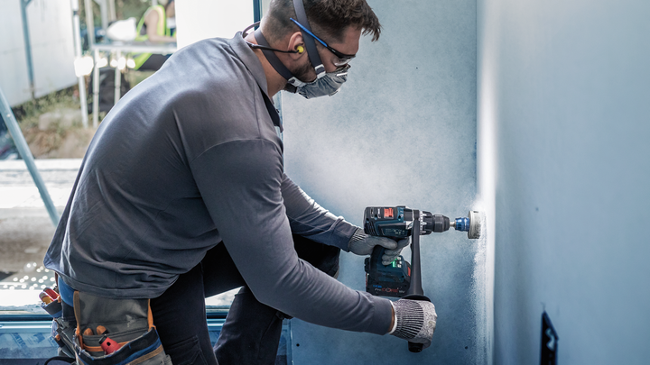 Worker wearing safety equipment drills a hole in drywall using a power tool.