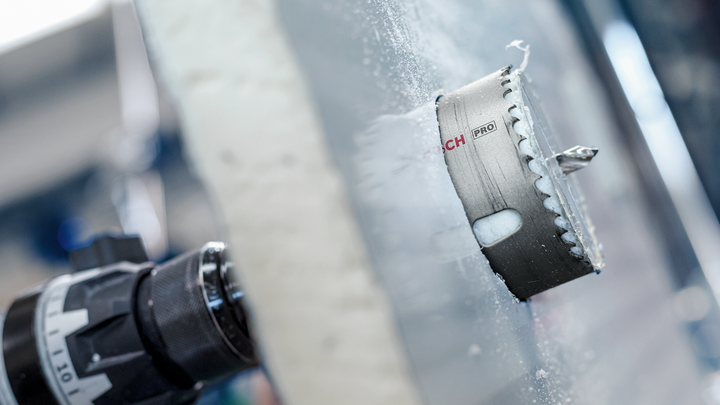 A hole saw attachment is drilling through a thick sheet of frosted glass.