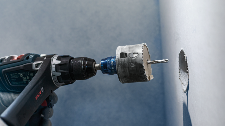 Worker wearing safety equipment drills a circular hole in a wall using a hole saw attachment.