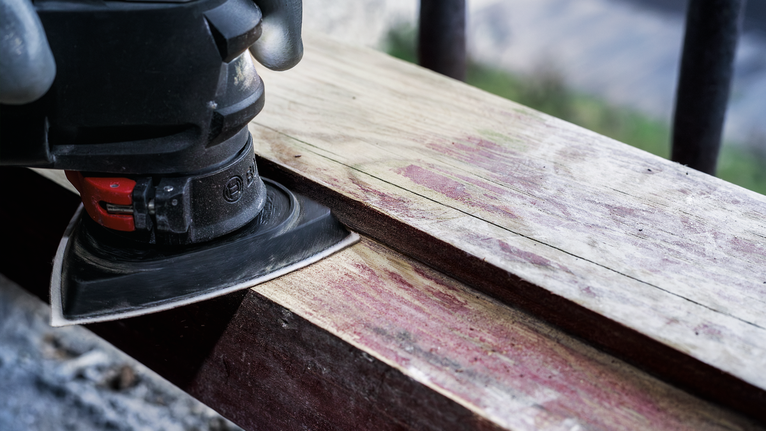 Person wearing safety equipment sands a wooden plank with a power sander.