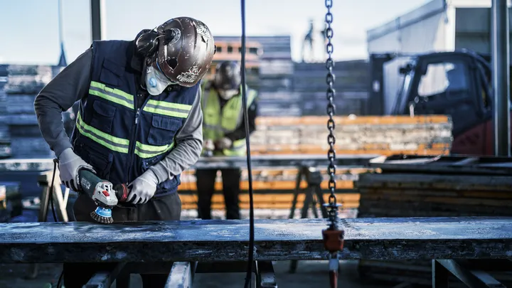 Object #200456904: 3089943_app_web_1 A person wearing safety equipment grinds a metal beam at a construction site.