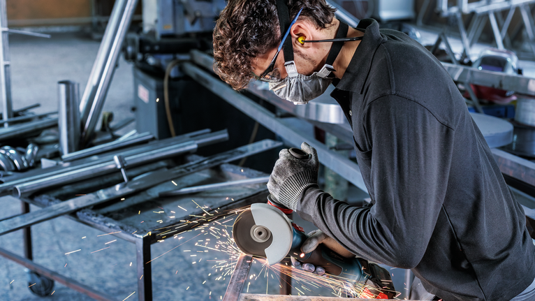Person wearing safety equipment uses an angle grinder to cut metal, producing sparks.