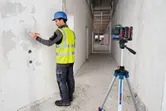 A person wearing safety equipment marks a wall guided by a laser leveling tool on a tripod.