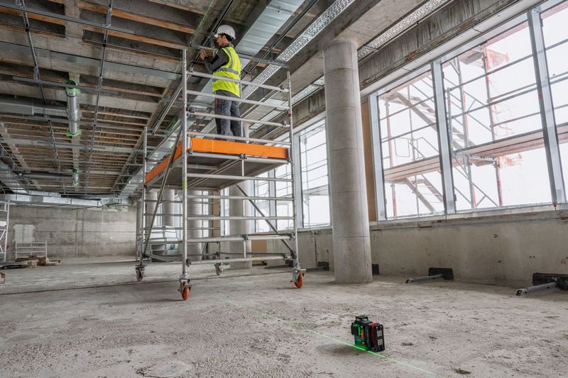 GLL 18V-120-33 CG A person wearing safety equipment works on scaffolding, with a laser leveling tool projecting lines.