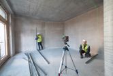 Two people wearing safety equipment align metal studs using a laser leveling tool in an unfinished room.