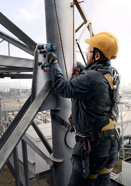 A person wearing safety equipment uses a cordless drill on a metal structure.