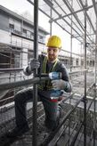 A person wearing safety equipment tightens a pipe joint with a cordless impact wrench on scaffolding.