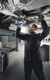 A person wearing safety equipment uses a power drill to work underneath a raised vehicle.