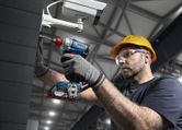 A person wearing safety equipment uses a cordless drill to install a security camera.