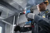 A person wearing safety equipment uses a cordless drill to fasten a metal duct.