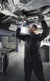 A person wearing safety equipment uses a cordless drill under a lifted vehicle.